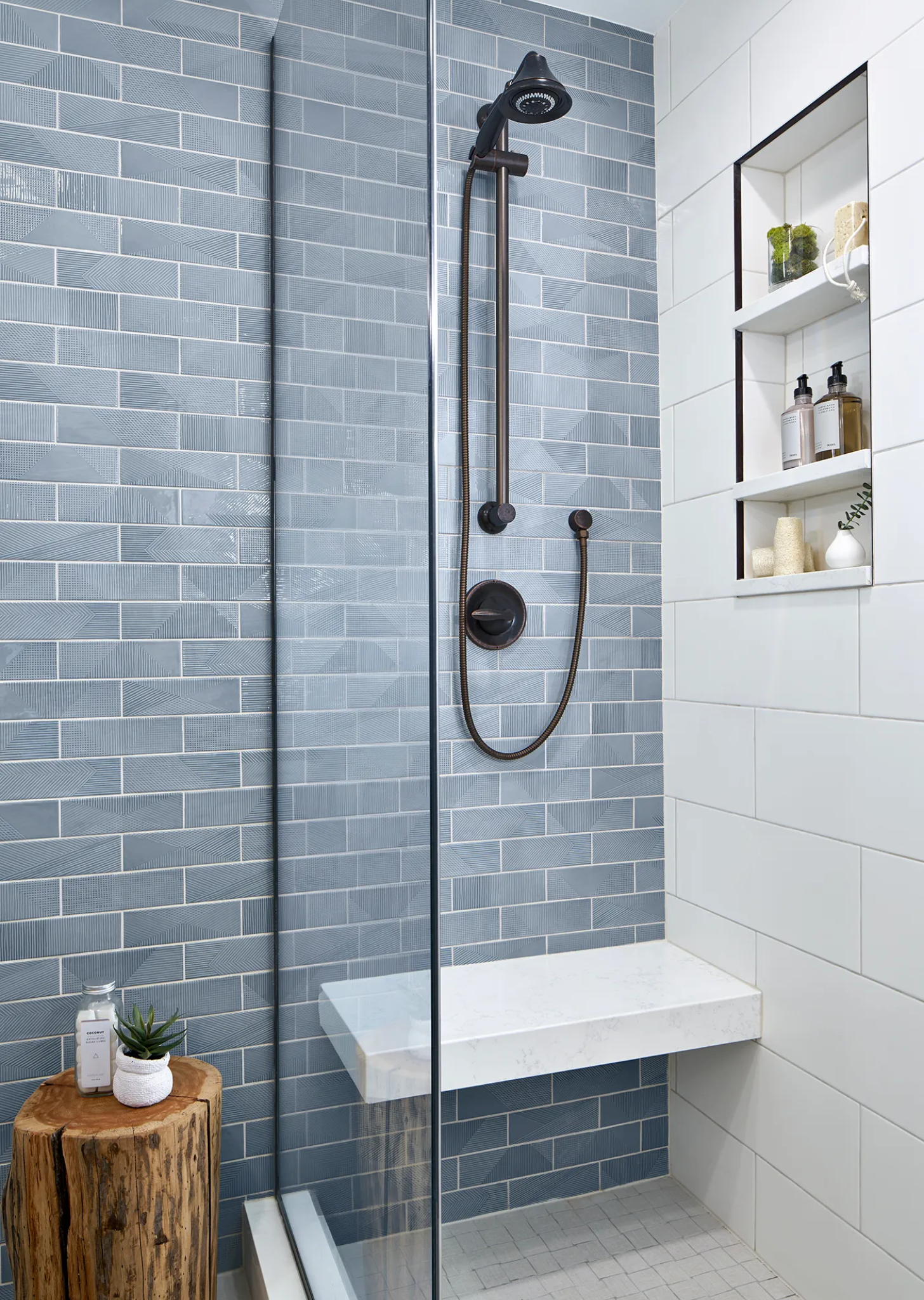 Contemporary bathroom with sleek glass shower, blue subway tiles, mounted showerhead, rustic wood stool, and recessed shelving for modern elegance.